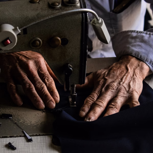 Manos ancianas de sastre cosiendo tela oscura en máquina de coser industrial, detalle del oficio artesanal
