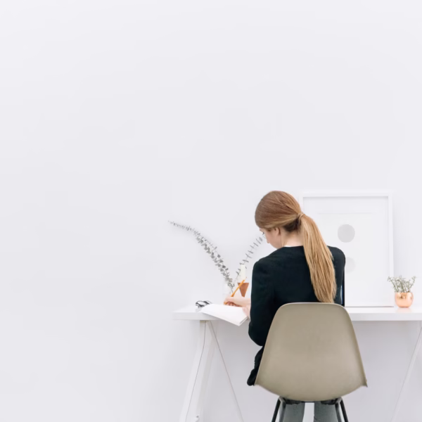 Espacio de trabajo minimalista con una persona de cabello largo recogido en coleta, sentada en una silla taupe frente a un escritorio blanco. Sobre el escritorio hay un cuaderno abierto, gafas, una bebida con pajilla, un cuadro con formas circulares, una rama seca en un florero transparente y una maceta pequeña de cobre con planta. El ambiente transmite simplicidad y enfoque.